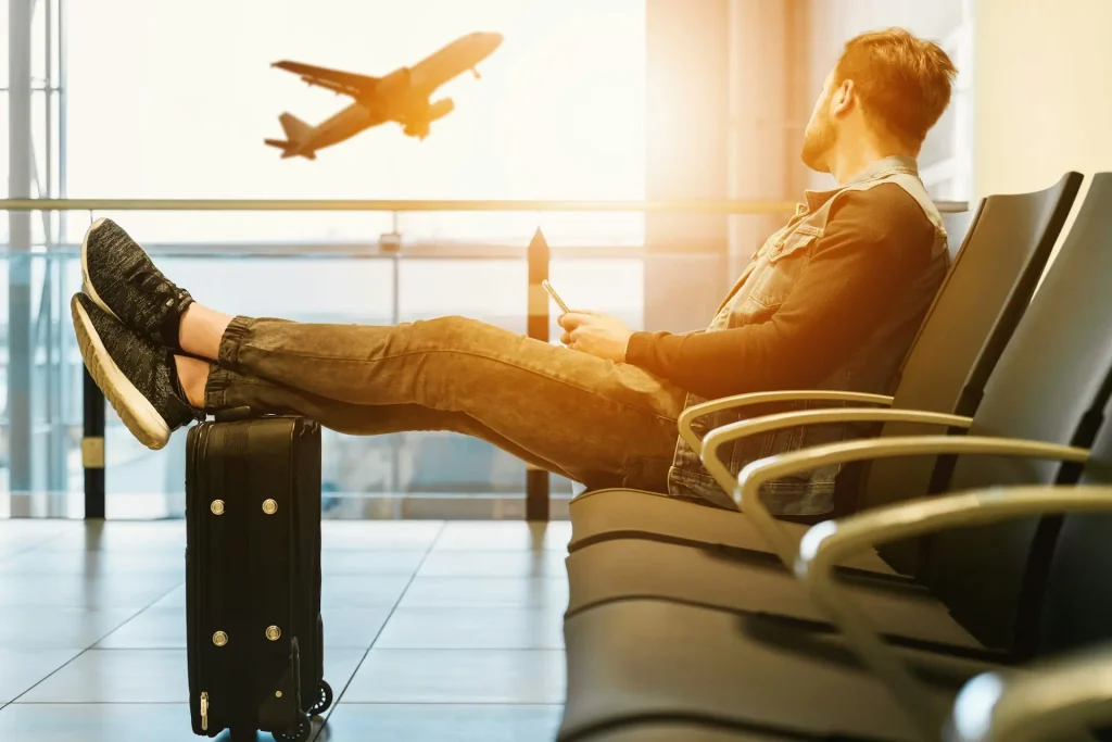 man waiting at airport with legs propped on suitcase watching plane take off through window