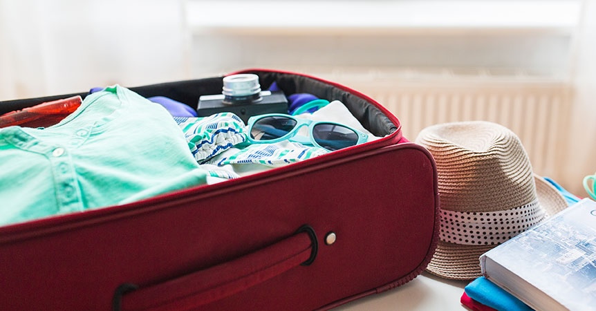 red carry on suitcase with clothes and sunglasses next to straw hat